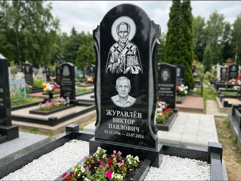 Памятник с гравировкой иконы: стела с образом в полный рост на обороте — гравировка образа Матроны Московской, низкие кусты спиреи в цвету, работа «Лебедева» в Ставрополе